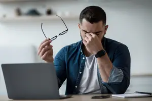 A man in a denim shirt holding a pair of glasses in front of his face while sitting at a desk with a laptop and notebook