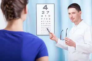 A man in a white lab coat stands in front of a woman wearing a blue t-shirt and holding a pen while looking at an eye chart with numbers and letters on a blue wall.