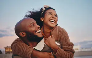 A man and woman are laughing and smiling while the man is carrying the woman on his shoulders on a beach.