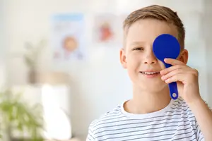 A boy wearing a striped shirt is holding a blue eye patch while smiling.
