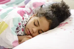 A young girl with curly hair sleeps peacefully on a bed with a colorful blanket.