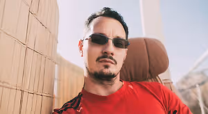 A man wearing sunglasses and a red t-shirt sits against a wooden wall, with mountains in the background and a blue sky overhead.