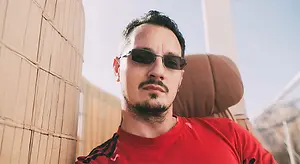 A man wearing sunglasses and a red t-shirt sits against a wooden wall, with mountains in the background and a blue sky overhead.