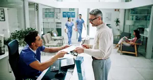 Two men are standing in front of a desk while one man looks at a paper. Behind them, two people are walking and a woman is sitting on a couch.