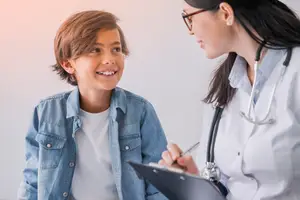 A smiling young boy in a denim jacket is standing next to a female doctor wearing glasses and a white coat, who is holding a clipboard and a pen, probably explaining something to the boy in a clinic room.