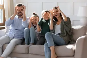 A family of four sitting on a couch, covering their eyes with their hands, smiling, and posing for a photo