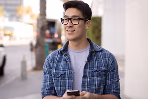 A man wearing glasses and a plaid shirt standing on the sidewalk looking at his phone