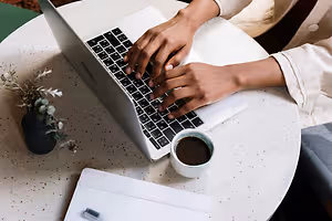 A person sitting on a chair while working on a laptop with a cup of coffee on the table