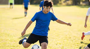 A boy wearing a blue shirt and goggles kicks a soccer ball on a field.