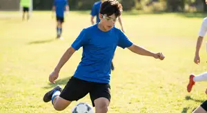 A boy wearing a blue shirt and goggles kicks a soccer ball on a field.