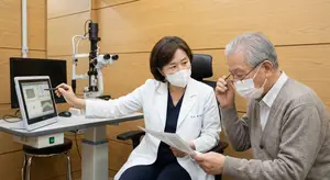 An elderly man with white hair is consulting with a doctor who is explaining something to him using a monitor in an eye clinic.
