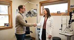A man and a woman in a white coat stand next to each other, shaking hands in a room with medical equipment.