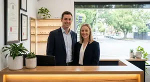 A man and a woman stand smiling in a professional setting, wearing business attire with potted plants and framed documents visible in the background.