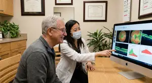 A man and woman sit in a medical office. The woman points at an eye scan on a computer screen.