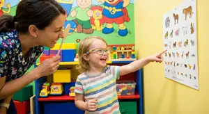 A young girl points to a picture of an animal on the wall while an adult female smiles and holds a wooden spoon
