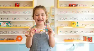 A young girl holds up a pair of pink and yellow glasses in a brightly lit room with shelves of colorful glasses in the background.