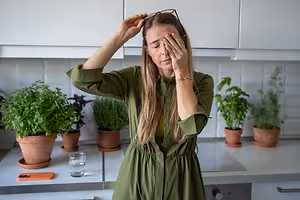 A woman in a kitchen suffering from a headache while closing her eyes