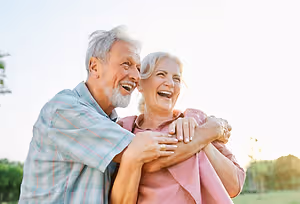An elderly couple, an older man and woman, are standing in a field, embracing each other with happy faces under a bright sky