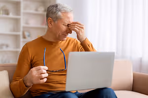 Elderly man sitting on a couch in his living room, looking at a laptop with his hand on his forehead.