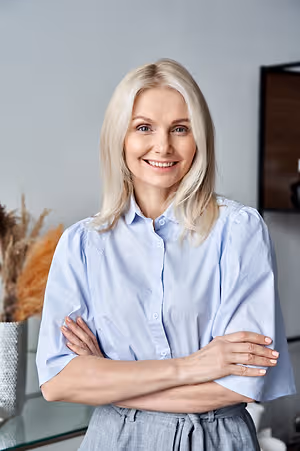 A woman with blonde hair is smiling and posing for a photo in an office room with a gray wall and a glass table.