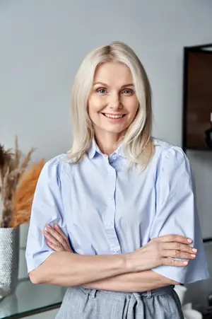 A woman with blonde hair is smiling and posing for a photo in an office room with a gray wall and a glass table.