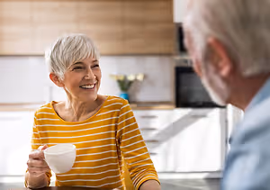 An older couple are sitting at a kitchen table and drinking coffee. The woman is smiling and holding a cup. The man is looking at her.