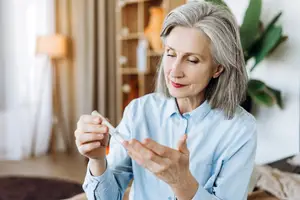 An elderly woman with gray hair holding a small object in her hands, perhaps a pill bottle, is sitting on a couch in a room.