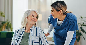 A nurse in blue scrubs is talking to an elderly woman in a striped shirt and green top in a living room with a couch, white curtains, and plants.