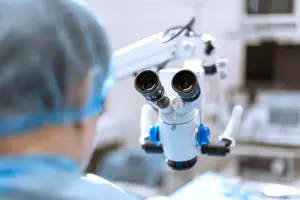 A person in blue medical attire and a hairnet looking through a microscope in a hospital room