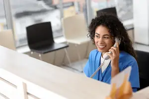 A woman wearing a blue uniform is sitting on a chair and holding a telephone while smiling in a hospital.
