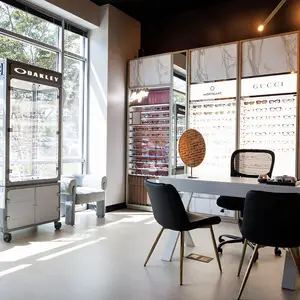 Inside an Oakley store, there are several display cases with sunglasses, a desk with two black chairs, a white chair, and a round display with a lamp on top. There are also several shelves with sunglasses on display, a round display case, and a wall with a white and black design.