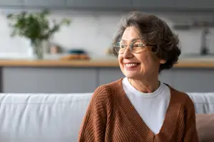 An older woman with short gray hair wearing glasses is smiling and looking at the camera while sitting on a couch in a living room.