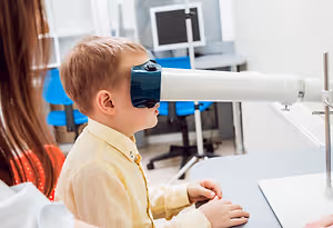 A young boy is looking through a microscope while a woman stands beside him in a room with a monitor and chairs