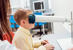 A young boy is looking through a microscope while a woman stands beside him in a room with a monitor and chairs