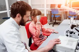 A man and a little girl are in a clinic. The man is looking at the girl while she is holding a phone in front of her face. Behind them are chairs and a table with a microscope. On the right side, there are some equipment.