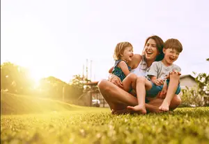 A woman is holding two kids in her lap, all of them are smiling in a grassy area