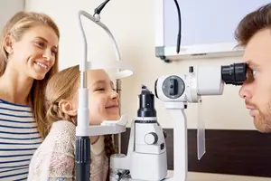 A young girl and her parents are looking through an ophthalmoscope while smiling at the camera.