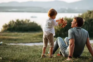 A child is standing on the grass in front of a man, both are holding wooden toys. The child is trying to throw the wooden toy to the man, and the man is trying to catch it. Behind them is a lake, mountains, and a boat. The area is surrounded by plants and trees, and there is a pathway on the side of the lake.