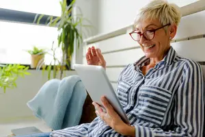 An elderly woman wearing glasses is sitting on a couch and using a tablet, smiling and waving