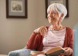 Senior woman with white hair wearing a pink shirt and glasses holds a book and looks towards a person in the room