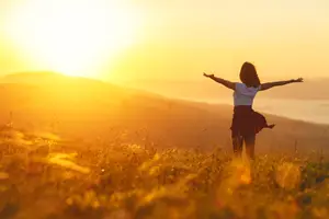 A woman standing on a grassy field with her arms wide open and the sun setting behind her