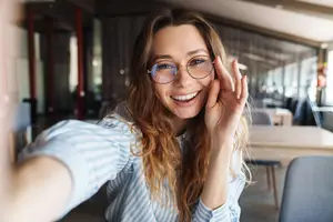 A woman wearing glasses is smiling and posing for a photo in an indoor space with chairs and tables.