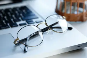 A pair of glasses with golden frames are placed on a laptop keyboard, with a wooden basket and a small, blurry object in the background.