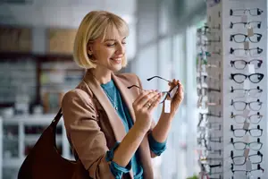 A woman holding glasses and smiling in front of a wall full of eyeglass frames.