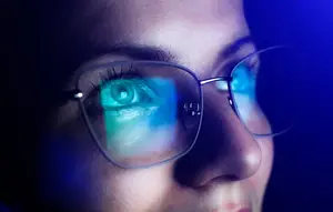 A woman with eyeglasses is wearing a blue light filter on her glasses, and her eyes are reflecting the blue light. She has long eyelashes and is looking at something in front of her. The light from the blue filter is illuminating her face, and the light is reflecting on her glasses.