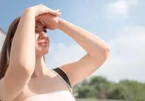 A woman with brown hair wearing a white sleeveless top and black strap on her shoulder is standing in an outdoor area and seems to be looking at something with a serious expression. Her right hand is on her forehead, and her left hand is on her shoulder. She is probably in a park or a garden, with trees and a blue sky with clouds in the background.