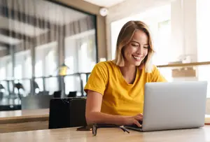 A woman sitting in front of a table with a laptop, smiling while looking at the laptop