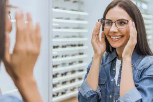 A woman in a denim jacket is smiling at herself in the mirror while wearing glasses