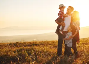 A family of four standing in a field during sunset, with a man holding a young child in the foreground and a young girl standing beside them
