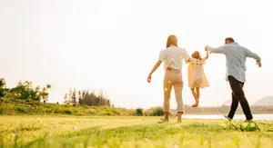 A family of three, a man, a woman, and a child, walking together on a grassy field near a body of water during sunset.
